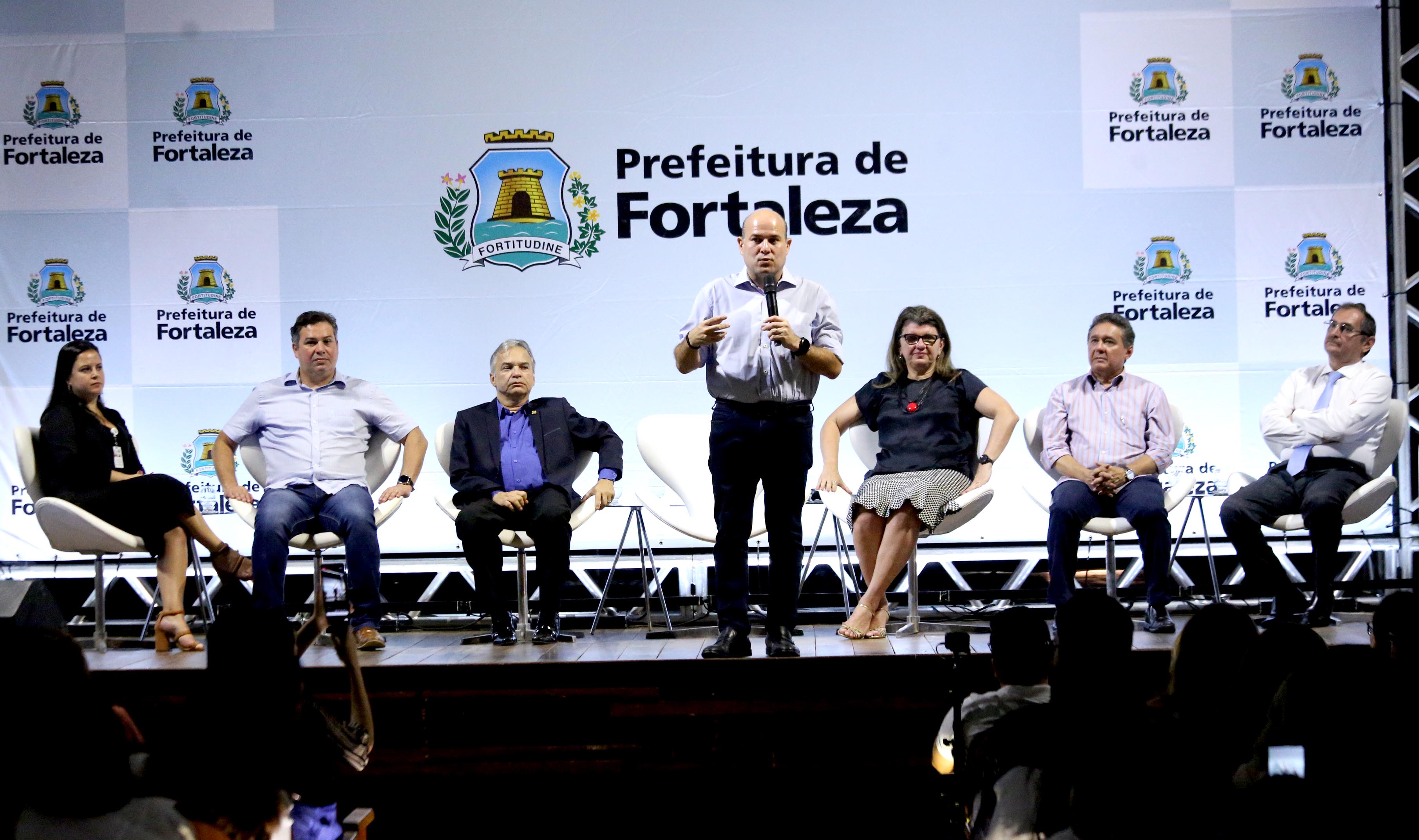 prefeito em cima de palco falando ao microfone e pessoas sentadas atrás